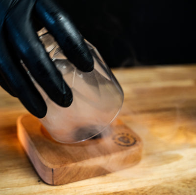 Black-gloved hand infusing a cocktail glass with smoke on a Helius Originals wooden board