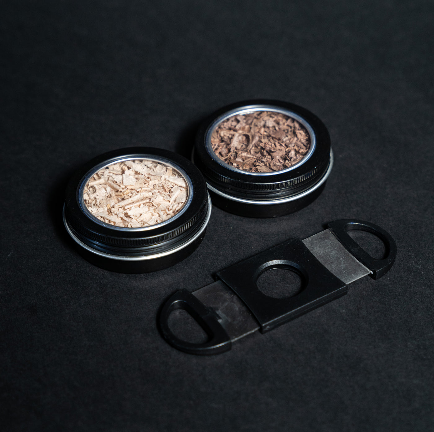 Helius Originals cocktail smoking accessories with two tins of premium wood shavings and a black cigar cutter on dark background