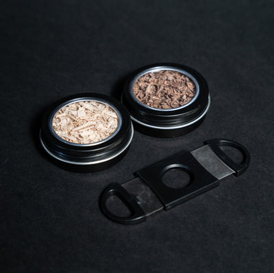 Helius Originals cocktail smoking accessories with two tins of premium wood shavings and a black cigar cutter on dark background