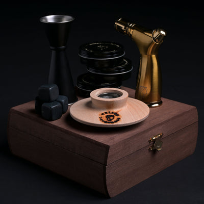 A collection of cocktail accessories displayed on a wooden box, including a cocktail chimney torch, wood shavings in tin containers, whiskey stones, a cocktail jigger, and a velvet bag.