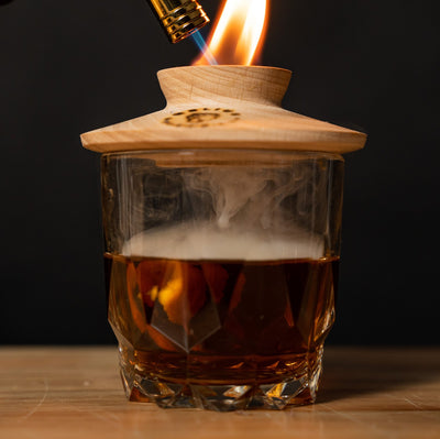 Luxury cocktail smoker in use with torch, infusing a whiskey glass with aromatic smoke using FSC-certified wood shavings