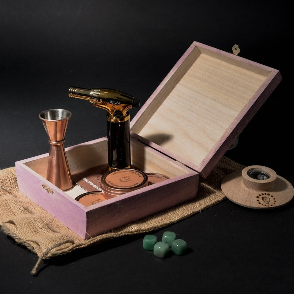 Helius Originals pink wood cocktail smoking kit with gold torch, jigger, wood shavings, and burlap bag in an open wooden box.