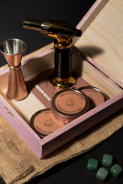 Smoke cocktail kit in open wood box with gold torch, copper jigger, wood shavings tins, recipe book, and whiskey stones.