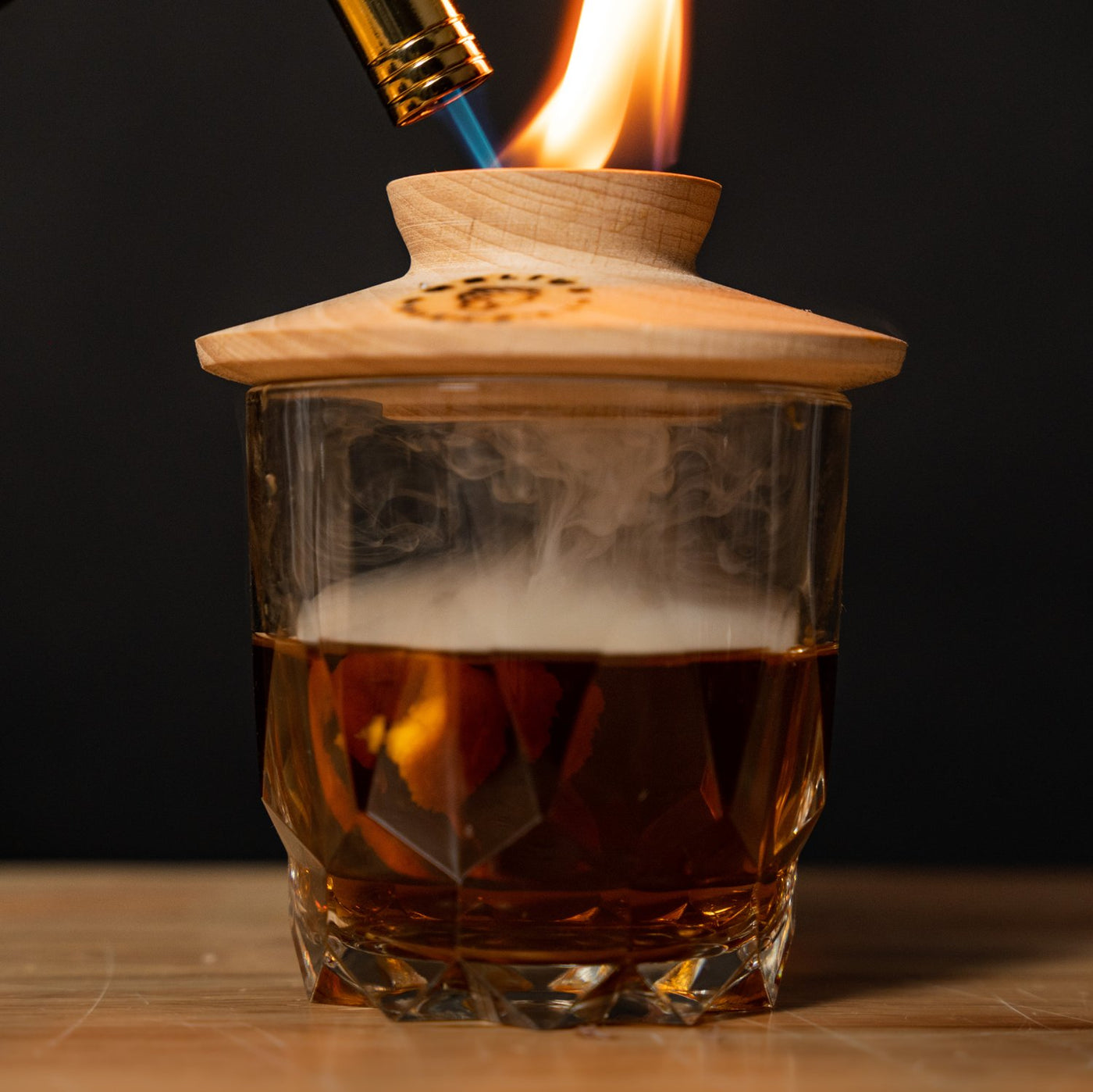 Whiskey cocktail in glass with smoke infuser lid and torch flame, part of the Helius Originals smoked cocktail kit.