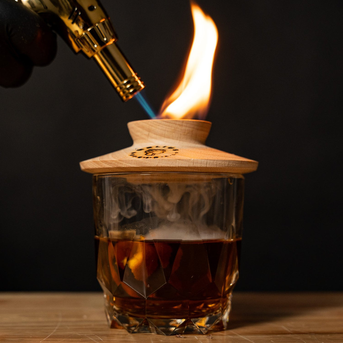 Smoked old fashioned cocktail being infused with flame using Helius Originals gold torch and wooden smoker lid, luxury barware for mixology and entertaining