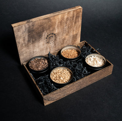 A set of wood shavings in four small tins, with one tin labeled 'Black Walnut', another labeled 'Cherry', a third labeled 'Maple', and a fourth labeled 'White Oak', presented in a wooden box with a burlap bag.